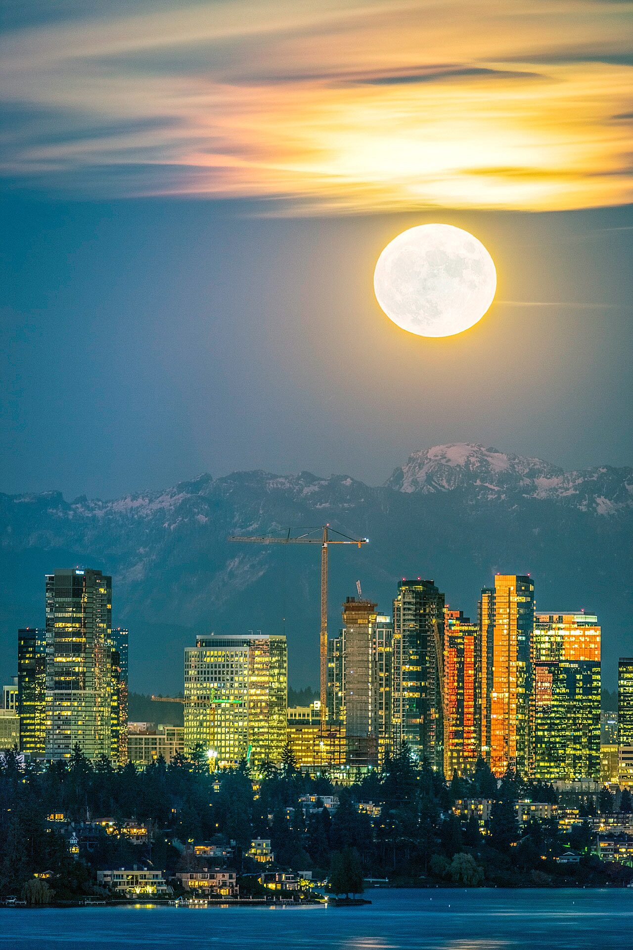 Landscape photo of moon-lit city with lake in foreground and mountains in background
