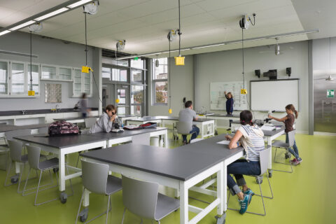 Interior photo of classroom with children sitting at tables and teacher pointing to whiteboard